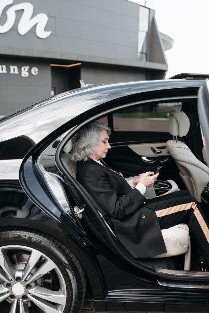 Senior woman in business attire sits in a luxury car, using a tablet outside a modern building.